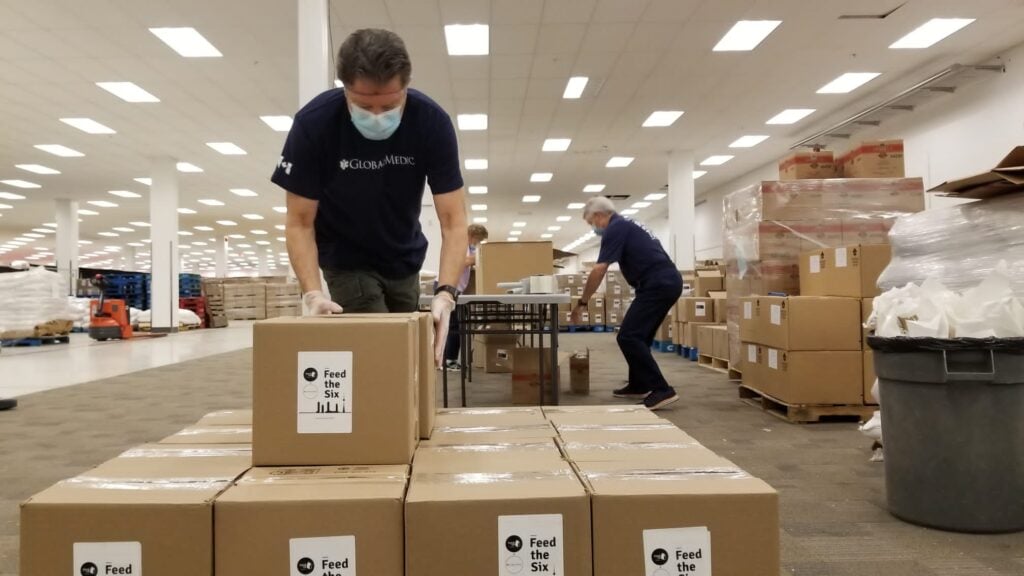 Volunteers stacking boxes onto a skid. The boxes have a sticker that says Feed the Six