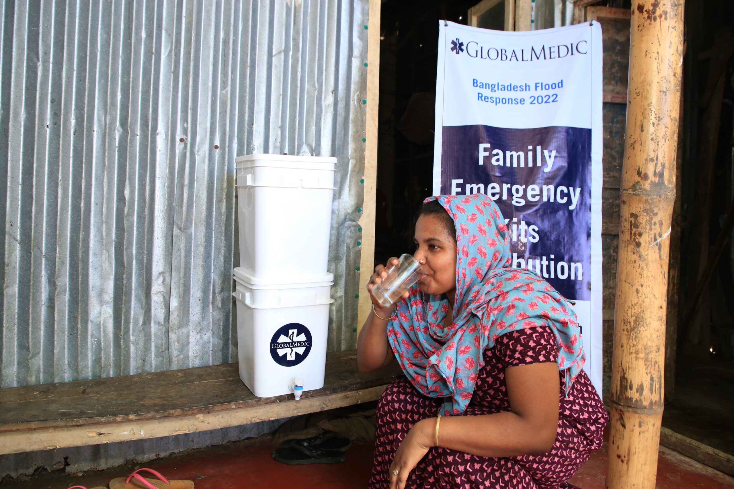 Sylhet Flooding Response: A woman drinking water from a glass kneeling in front of a water filtration unit unit